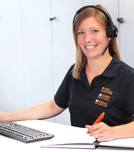 Eine Frau mit Headset und schwarzem Poloshirt sitzt lächelnd an einem Schreibtisch, tippt auf einer Tastatur und hält einen Stift über ein aufgeschlagenes Notizbuch.